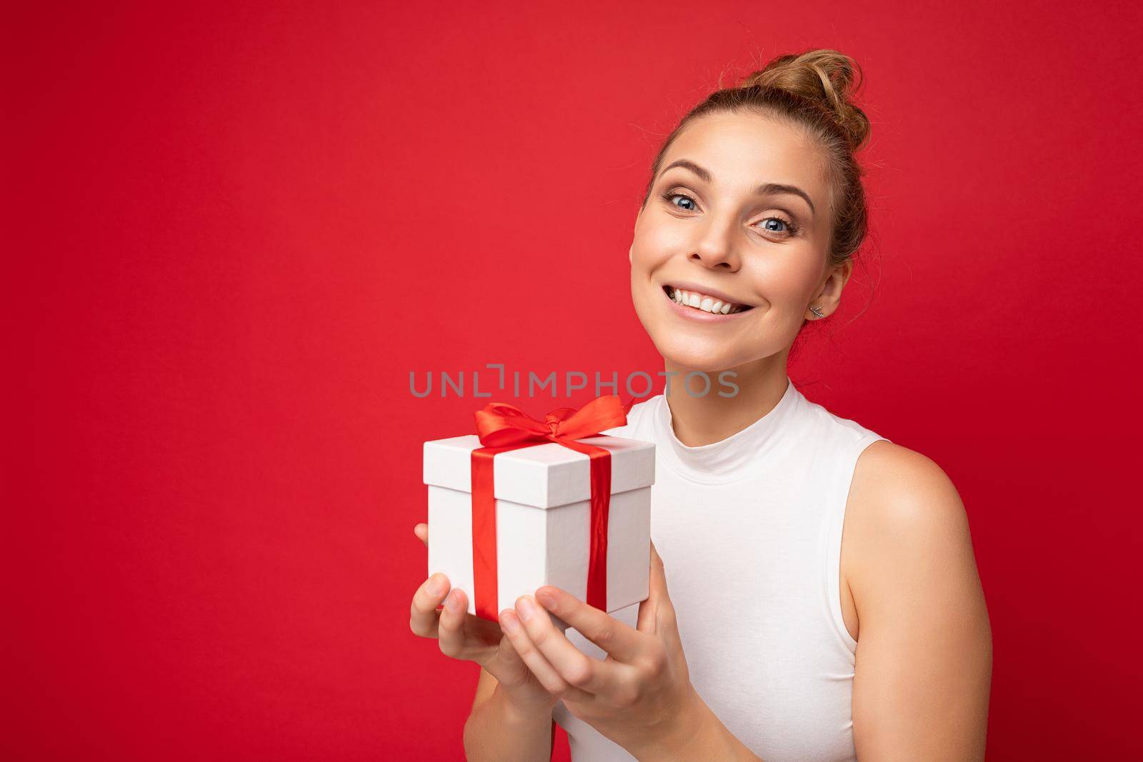Photo shot of beautiful attractive cute positive happu smiling young blonde woman isolated over red background wall with copy space for text wearing casual white top holding gift box with red ribbon. Surprise and present concept.
