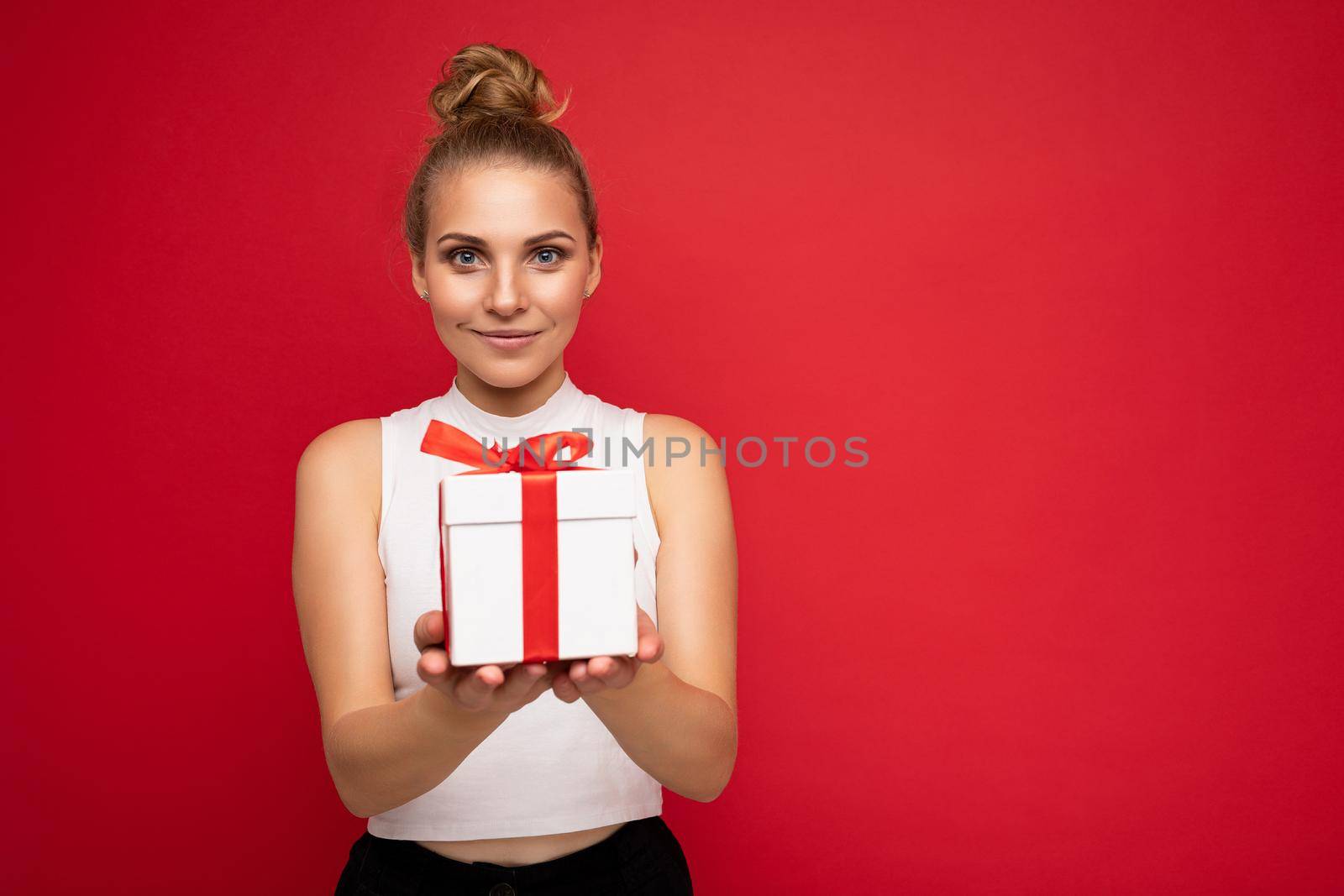 Photo shot of beautiful attractive cute positive young blonde woman isolated over red background wall with copy space for text wearing casual white top holding gift box with red ribbon. Birthday concept.