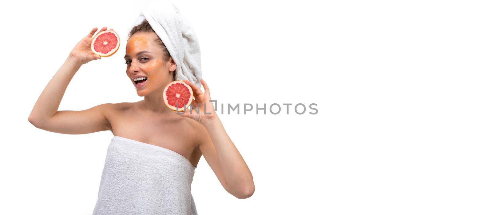 a woman after a shower with an orange cosmetic mask on her face is holding a cut grapefruit and dancing on a white background.