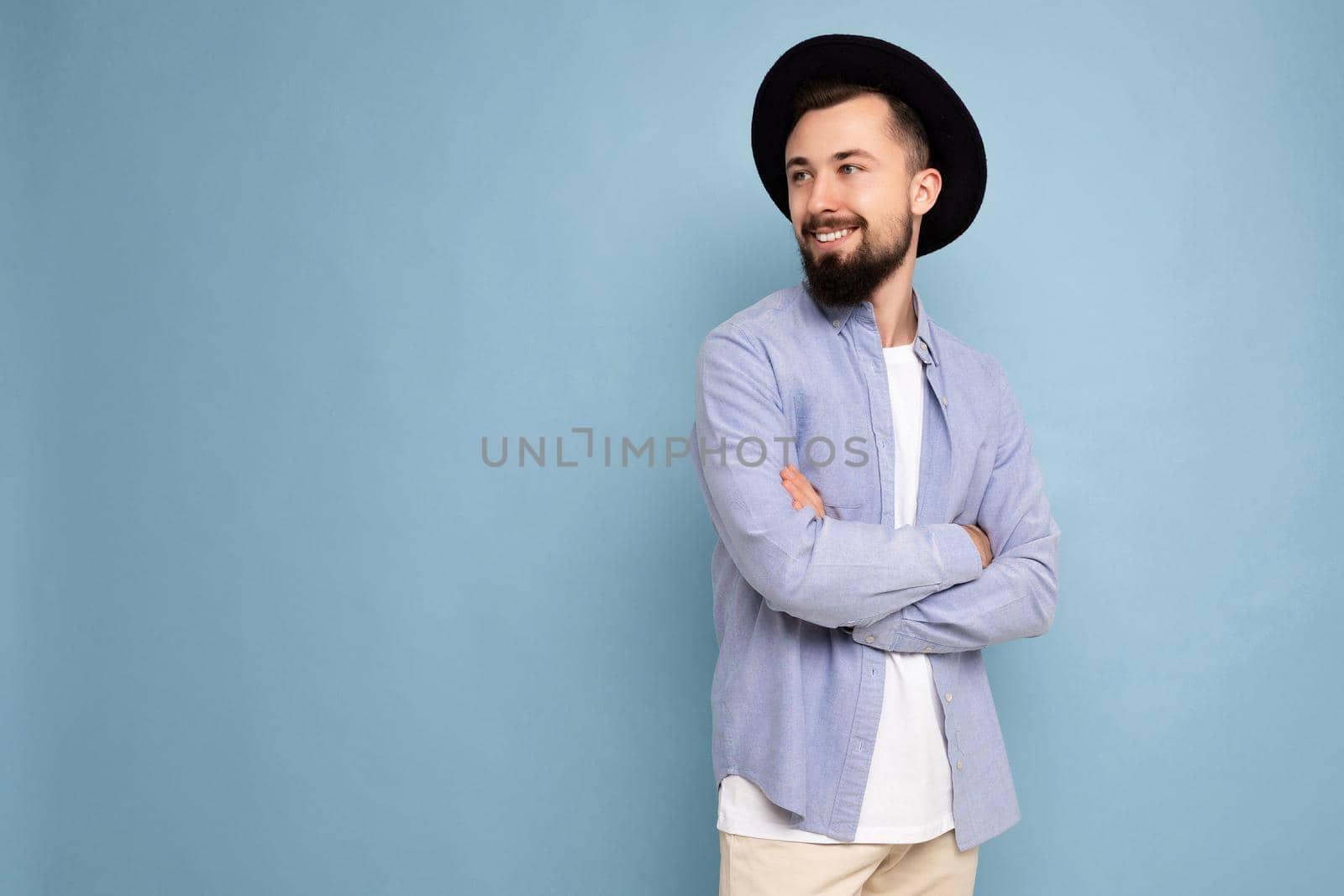 Photo shot of positive happy smiling good looking young brunet bearded man wearing casual blue shirt and white t-shirt and stylish black hat poising isolated on blue background with copy space for text.
