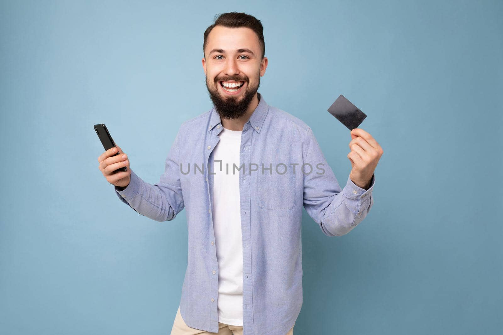 Photo of good looking attractive smiling brunet unshaven young man wearing casual blue shirt and white t-shirt isolated over blue background wall holding credit card and mobile phone.