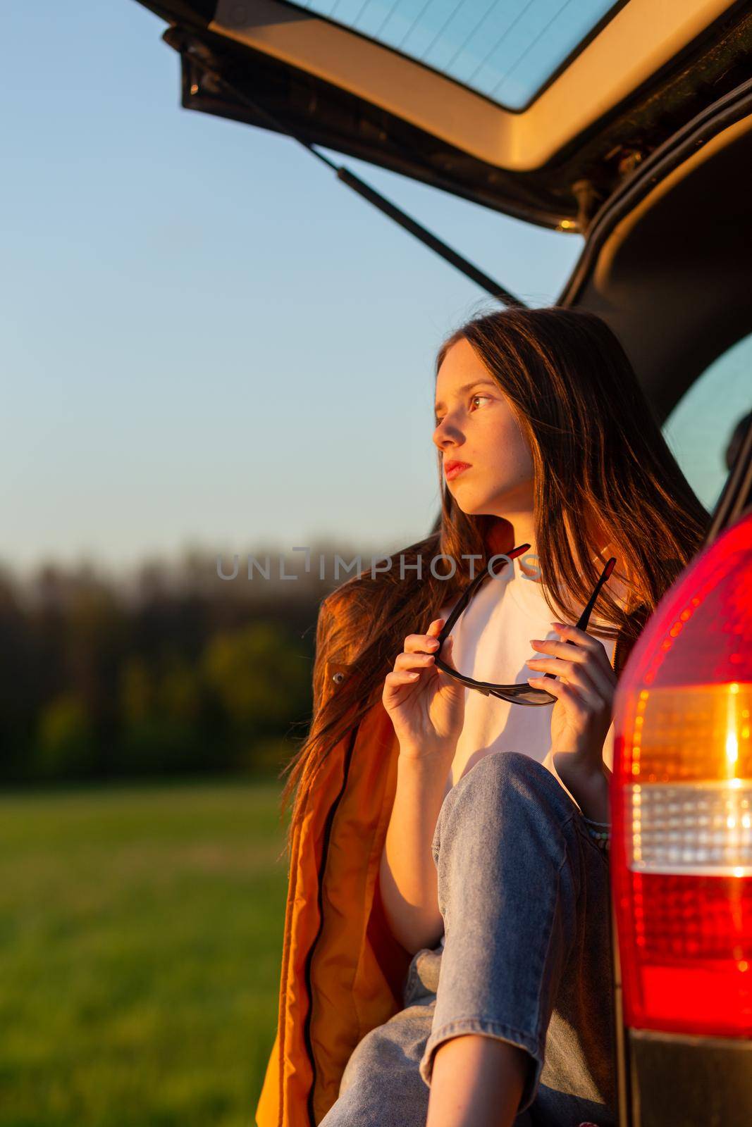 Pretty sad teenage girl with sun glasses sitting alone in a car trunk. Car travel concept.