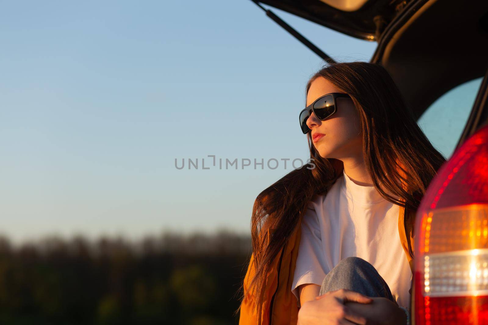 Pretty sad teenage girl with sun glasses sitting alone in a car trunk. Car travel concept.