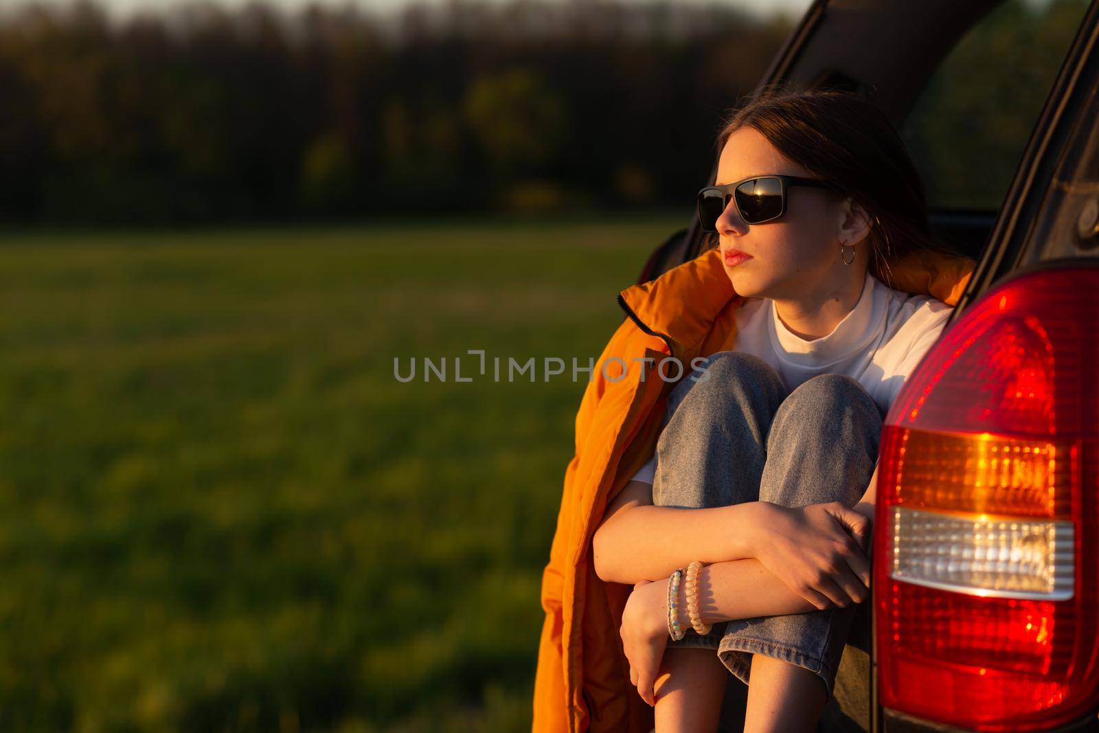 Pretty sad teenage girl with sun glasses sitting alone in a car trunk. Car travel concept.