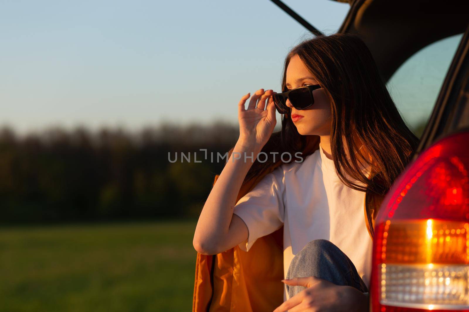 Pretty sad teenage girl with sun glasses sitting alone in a car trunk. Car travel concept.