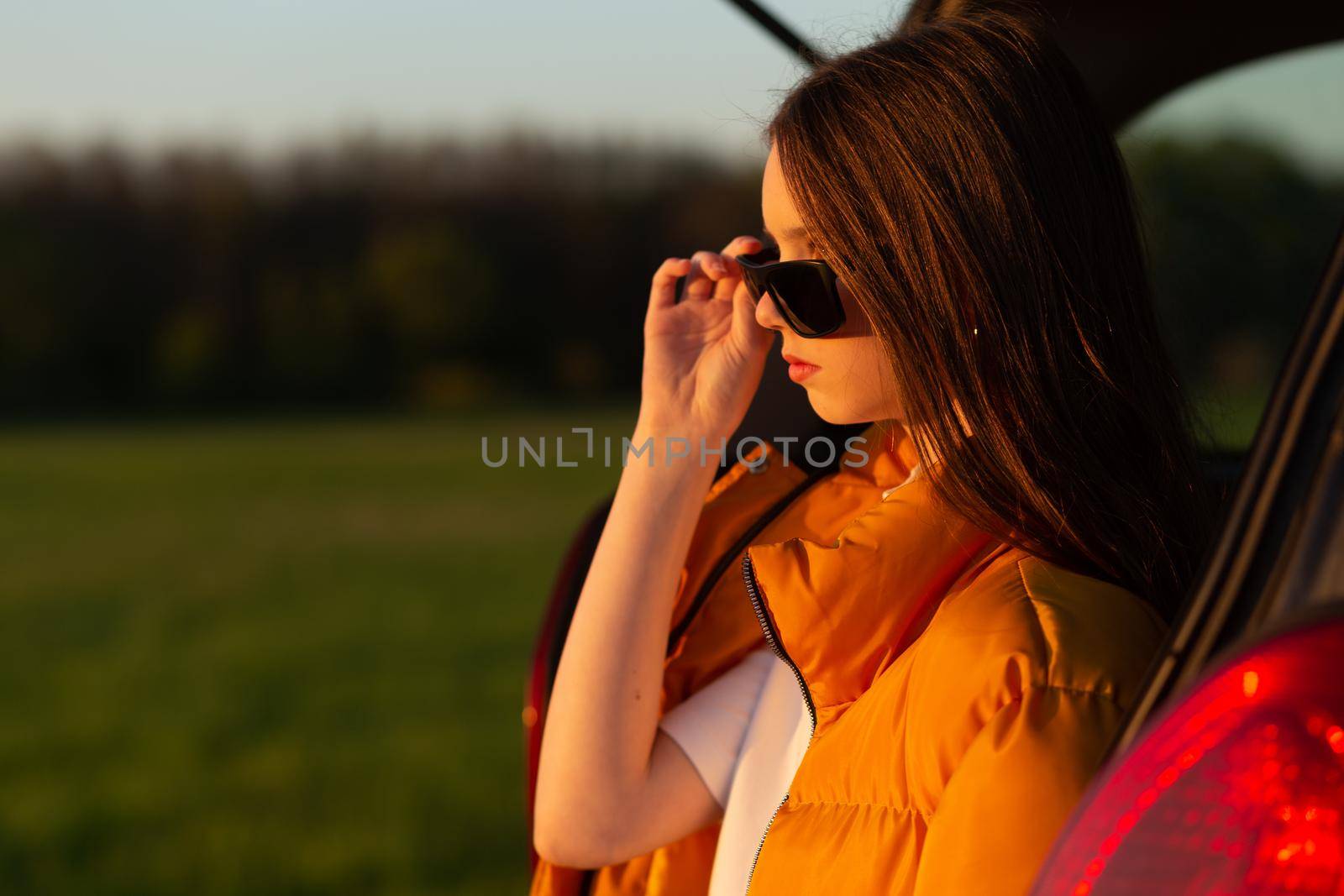 Pretty sad teenage girl with sun glasses sitting alone in a car trunk. Car travel concept.