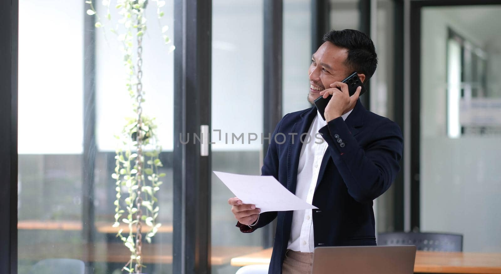 Asian businessman sitting on the phone with a customer with a laptop and document at his office desk. by wichayada