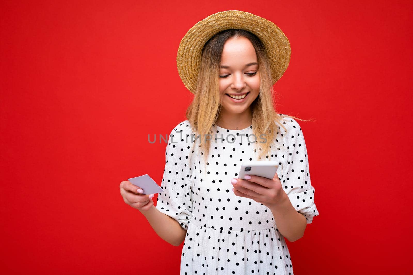 Photo of cheerful pretty blonde woman in summer outfit using mobile phone and holding credit card making payment online isolated over red background. Copy space by TRMK