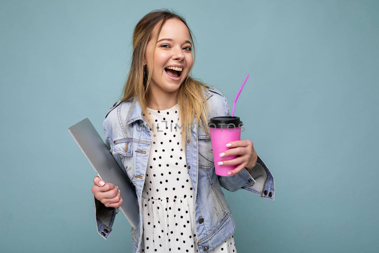 Photo of smiling happy young blonde woman wearingdenim jacket isolated over blue background, carrying laptop and drinking coffee. Copy space by TRMK