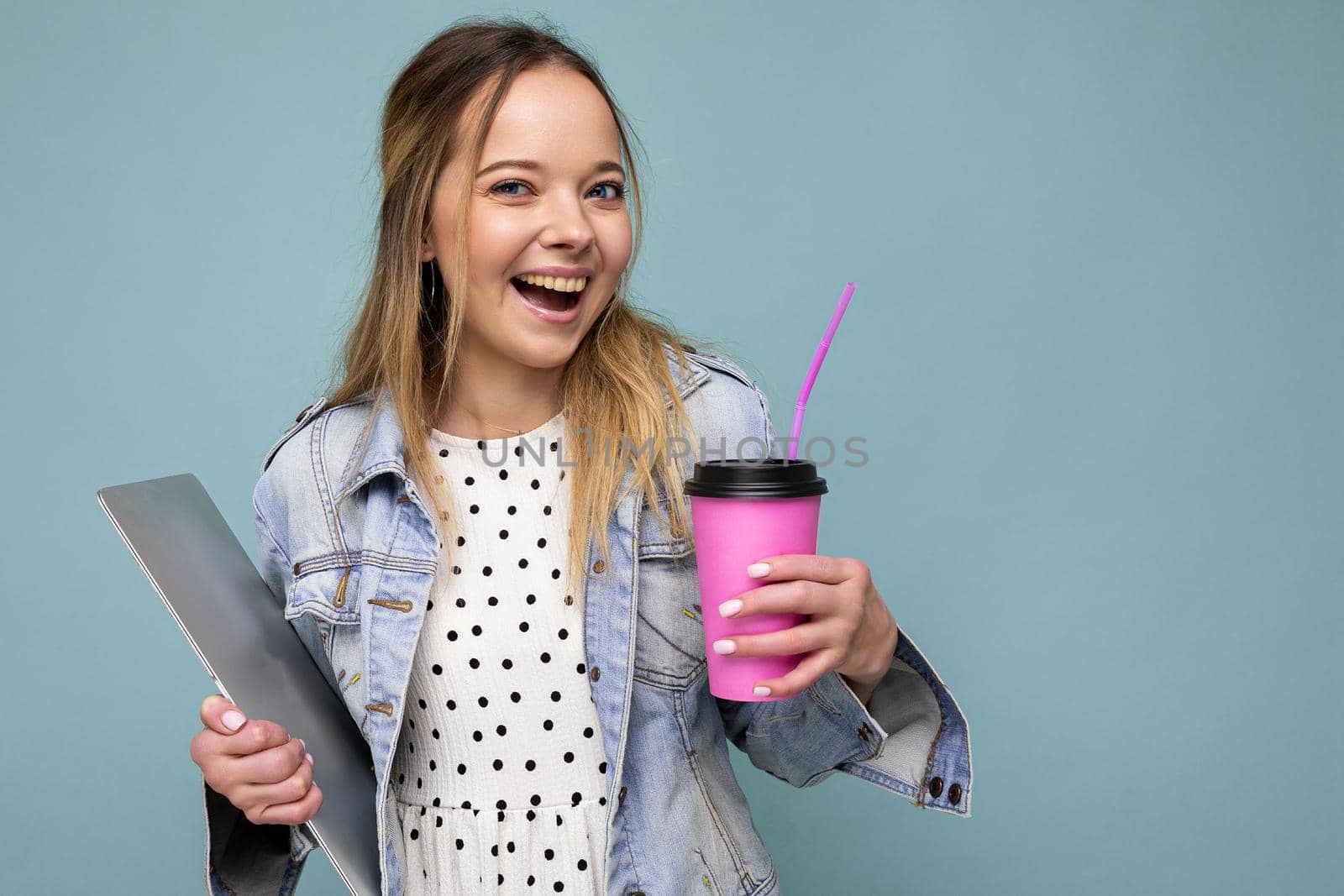 Image of joyful european blond woman smiling and standing against blue wall isolated with silver notebook and takeaway coffee in hands.