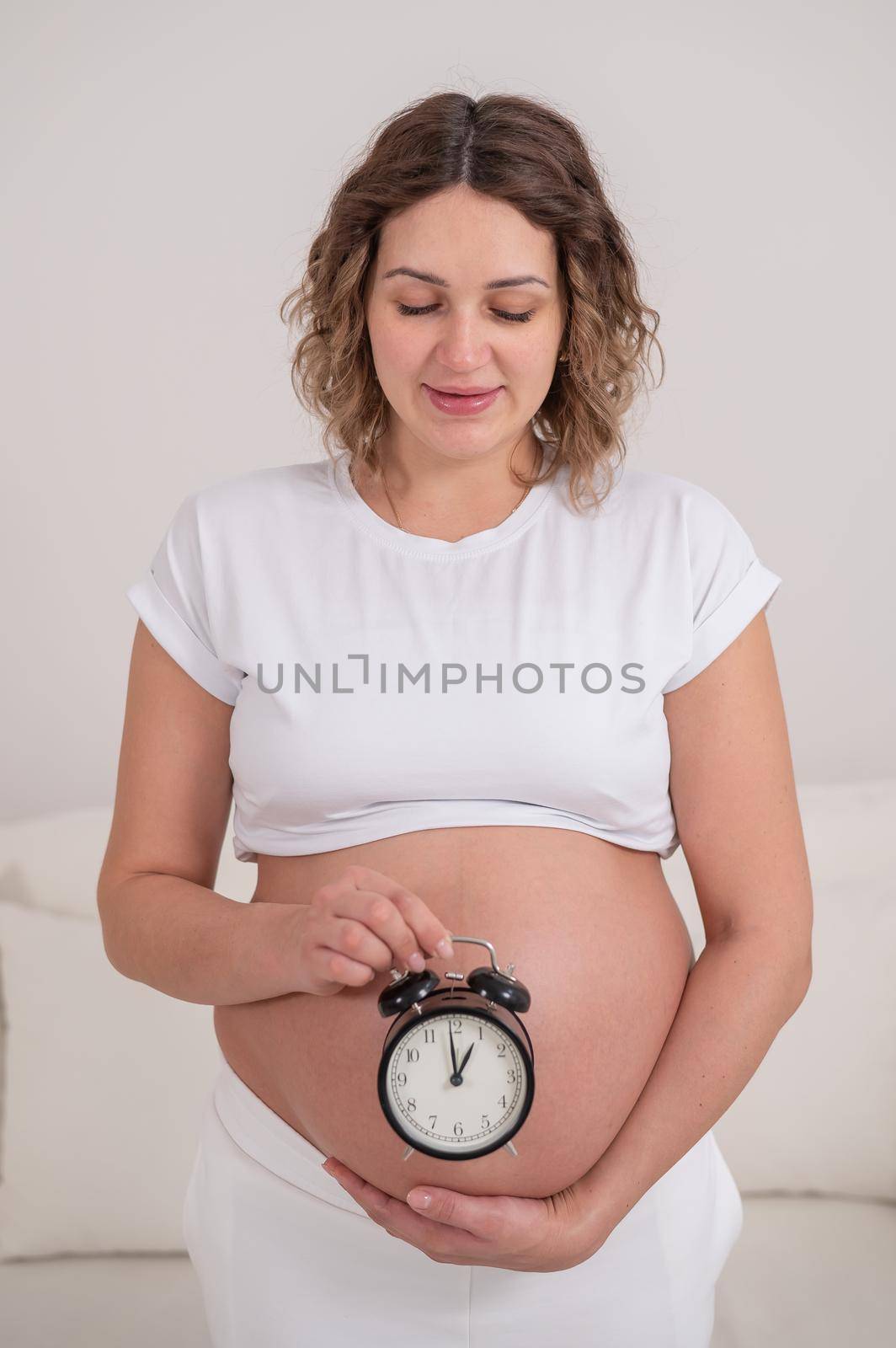 A pregnant woman with a naked belly holds an alarm clock. Intended date of birth