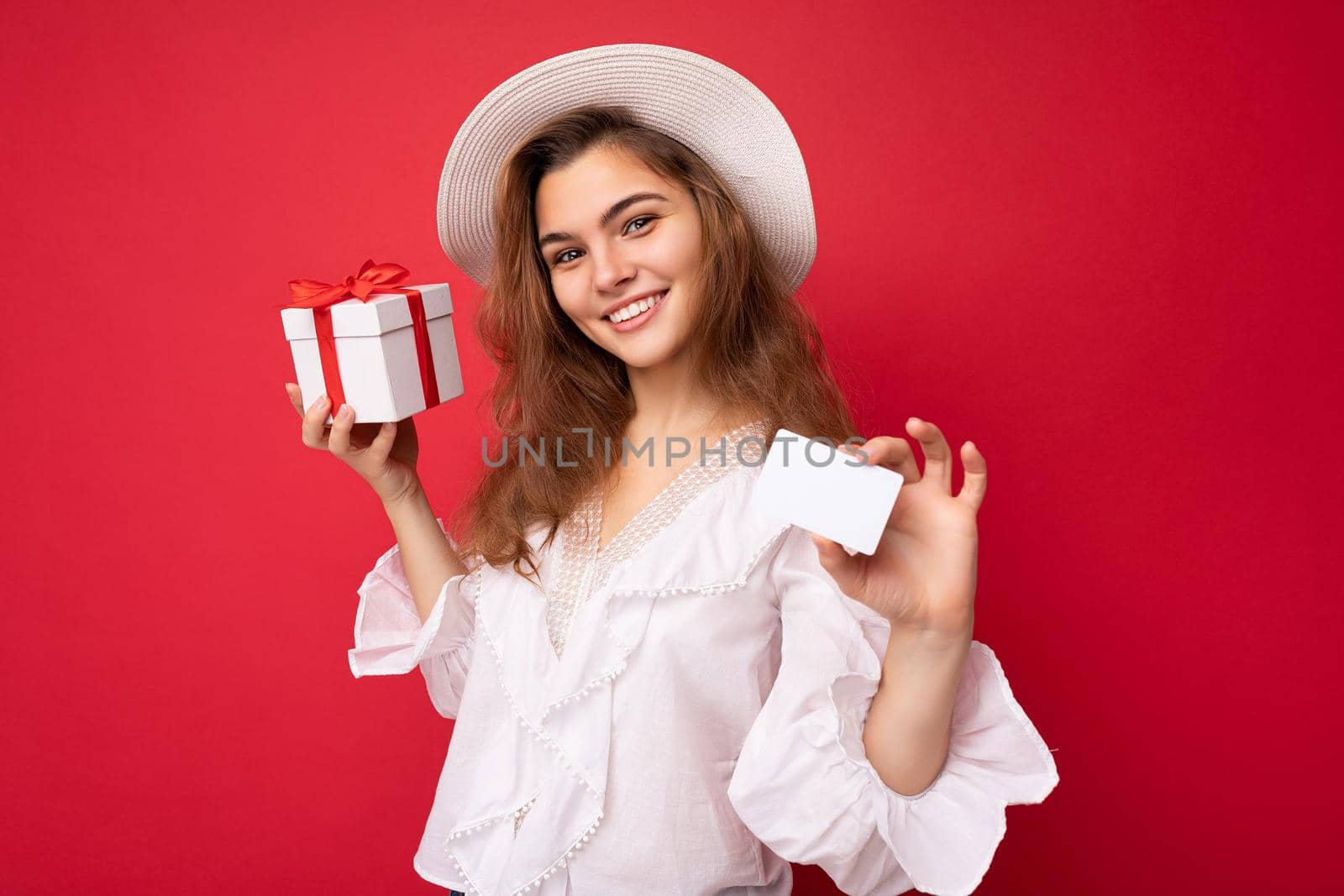 Portrait of young attractive caucasian hipster woman in trendy casual clothes and white hat. Sexy carefree female person posing isolated near red wall in studio. Positive model with natural makeup. Holding white gift box with red ribbon and credit card for shopping. Empty space.