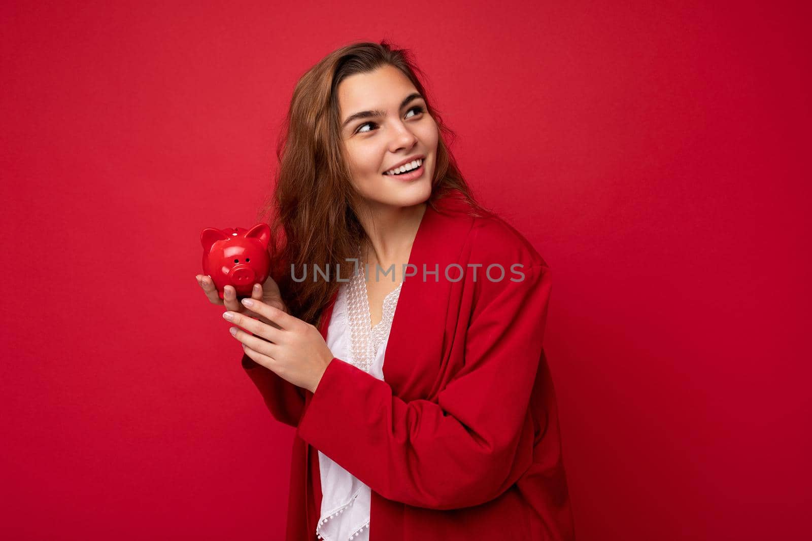 Young beautiful european stylish brunette woman wearing white blouse and red cardigan isolated over red background with positive sincere emotions. Holding little red moneybox. Free space by TRMK