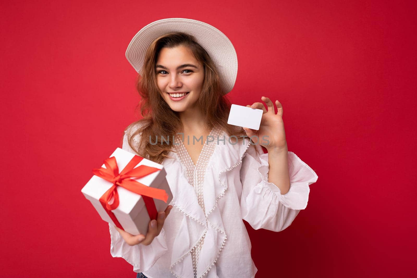 Young beautiful european stylish brunette woman wearing white blouse and fashionable hat isolated over red background with positive sincere emotions. Simple and natural looking at the camera. Holding gift box and credit card. Free space. Mock up by TRMK