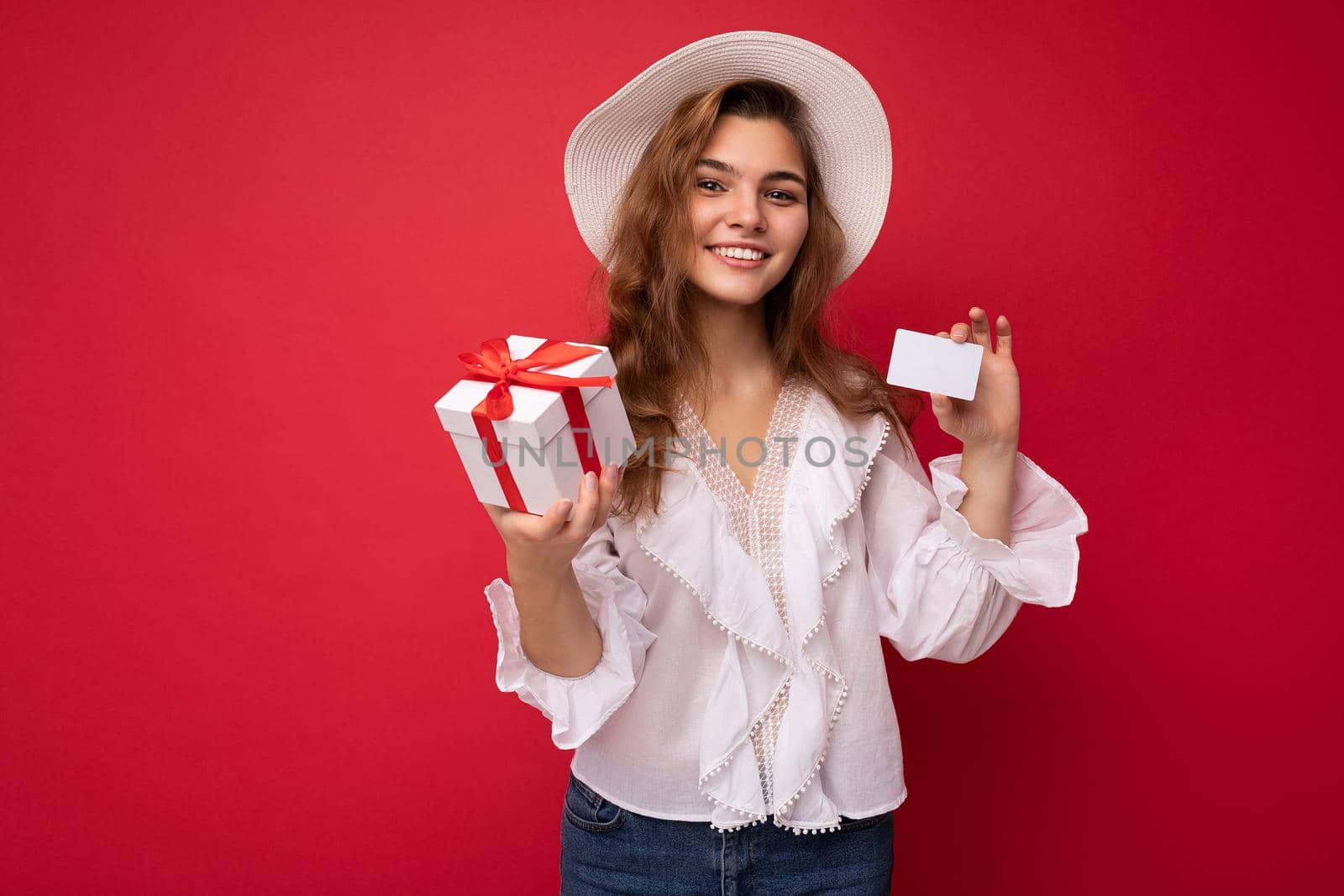Portrait of positive cheerful fashionable woman in formalwear holding gift box and credit card looking at camera isolated on red background with copy space.