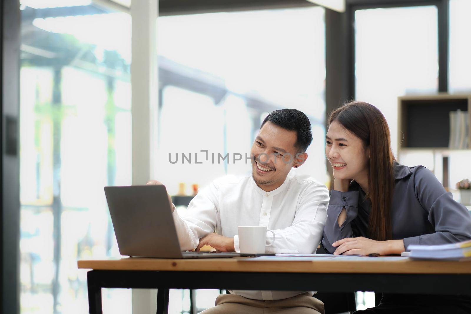 Two employees in a modern office, an Asian man and a woman working at a table, colleagues discussing and consulting, thinking about a joint project by wichayada