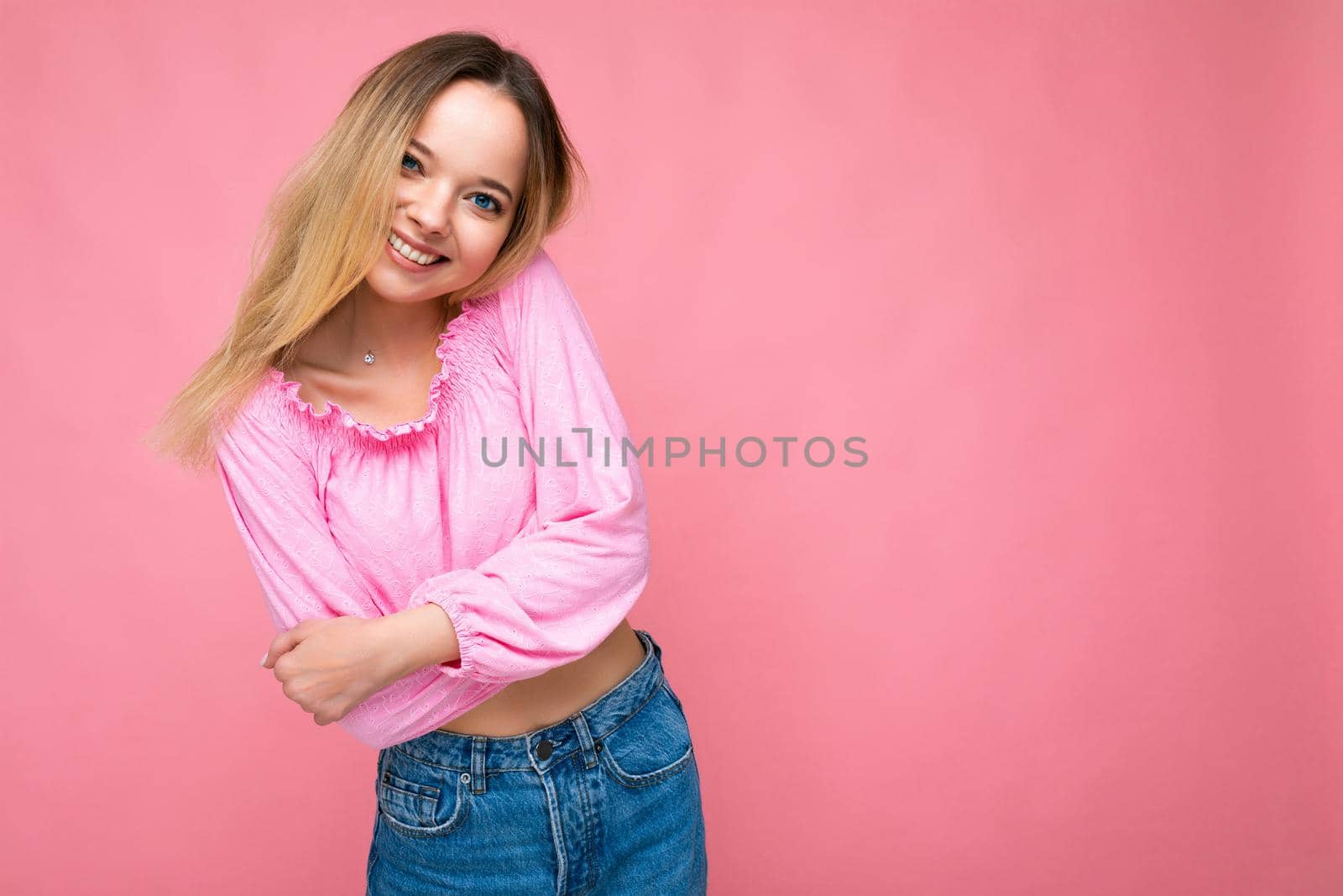 Portrait of young beautiful smiling hipster blonde woman in trendy pink crop top blouse. Sexy carefree female person posing isolated near pink wall in studio. Positive model with natural makeup. Copy space.