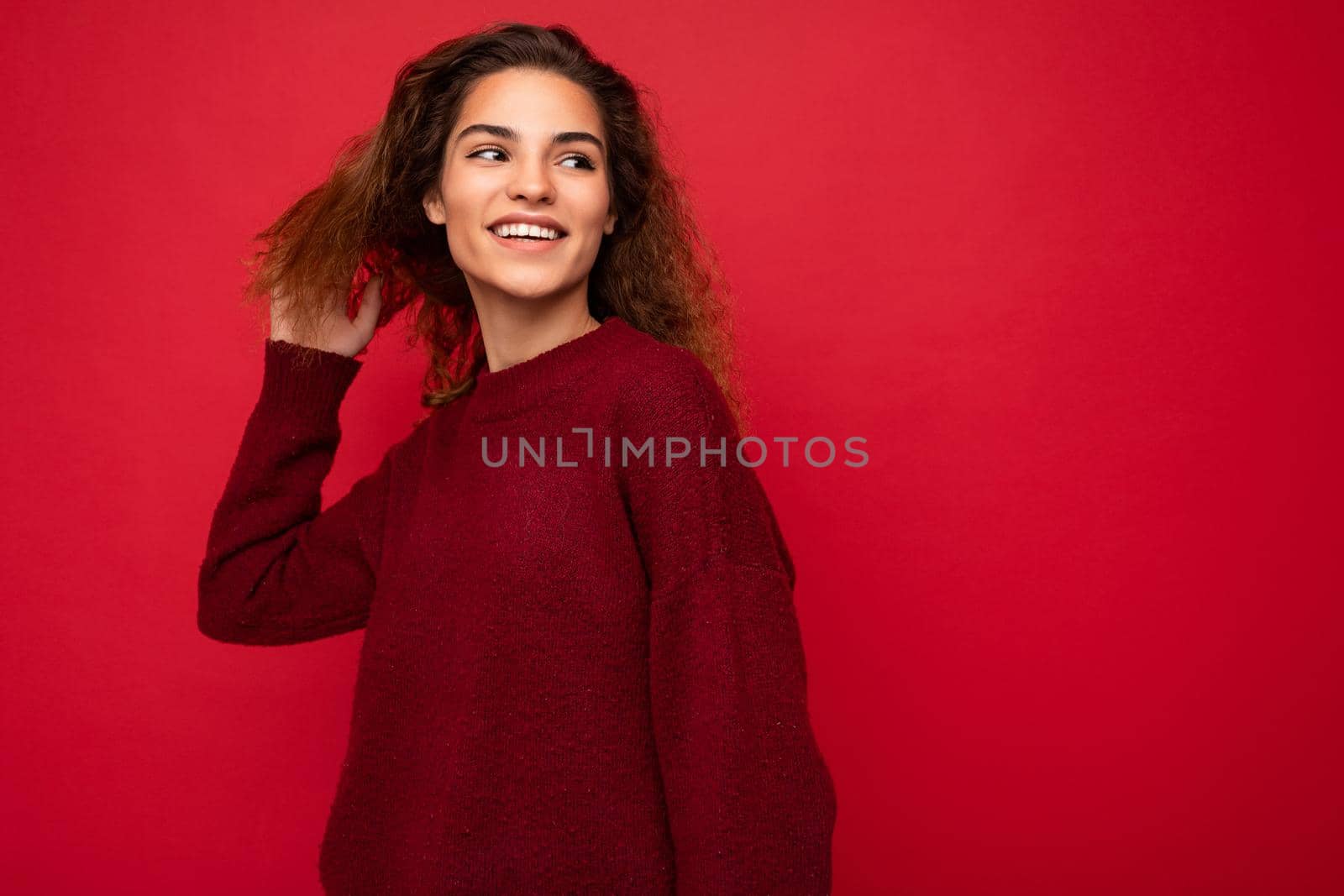 Photo of beautiful attractive charming young brunette curly woman wearing stylish dark red sweater isolated over red background wall with free space for text by TRMK