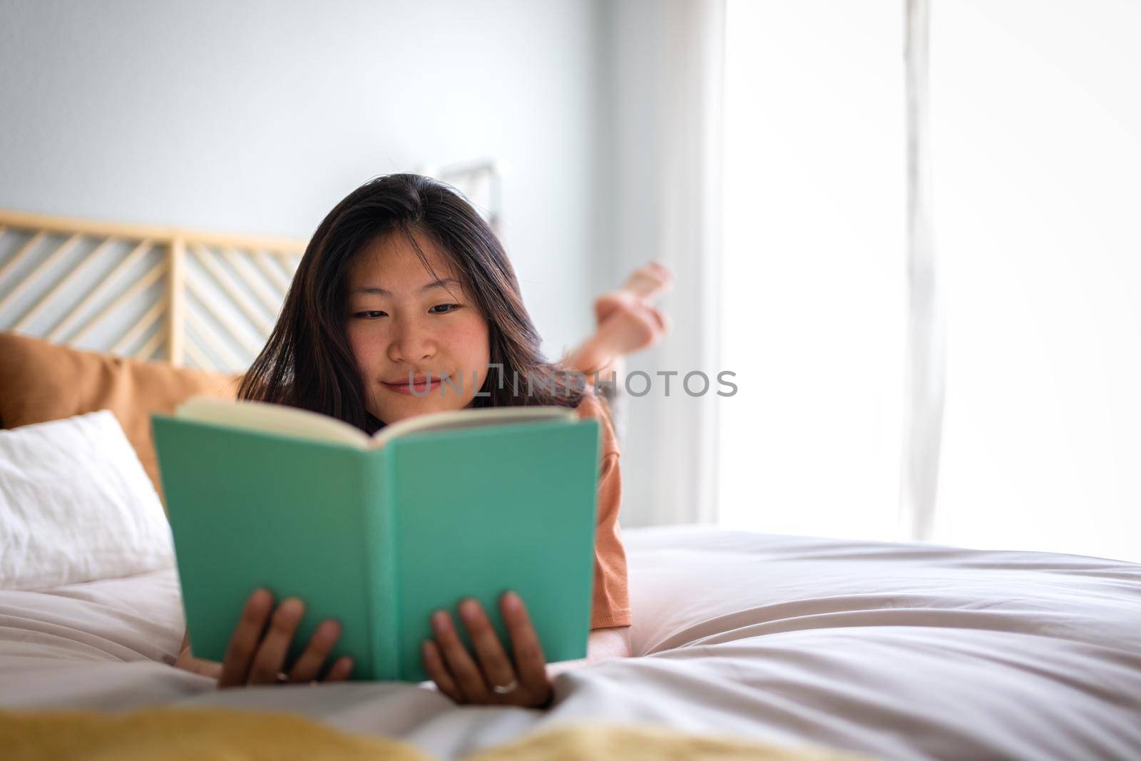 Teen asian college student reading book at home lying on bed in cozy room. Copy space. Comfort, leisure and home concepts.