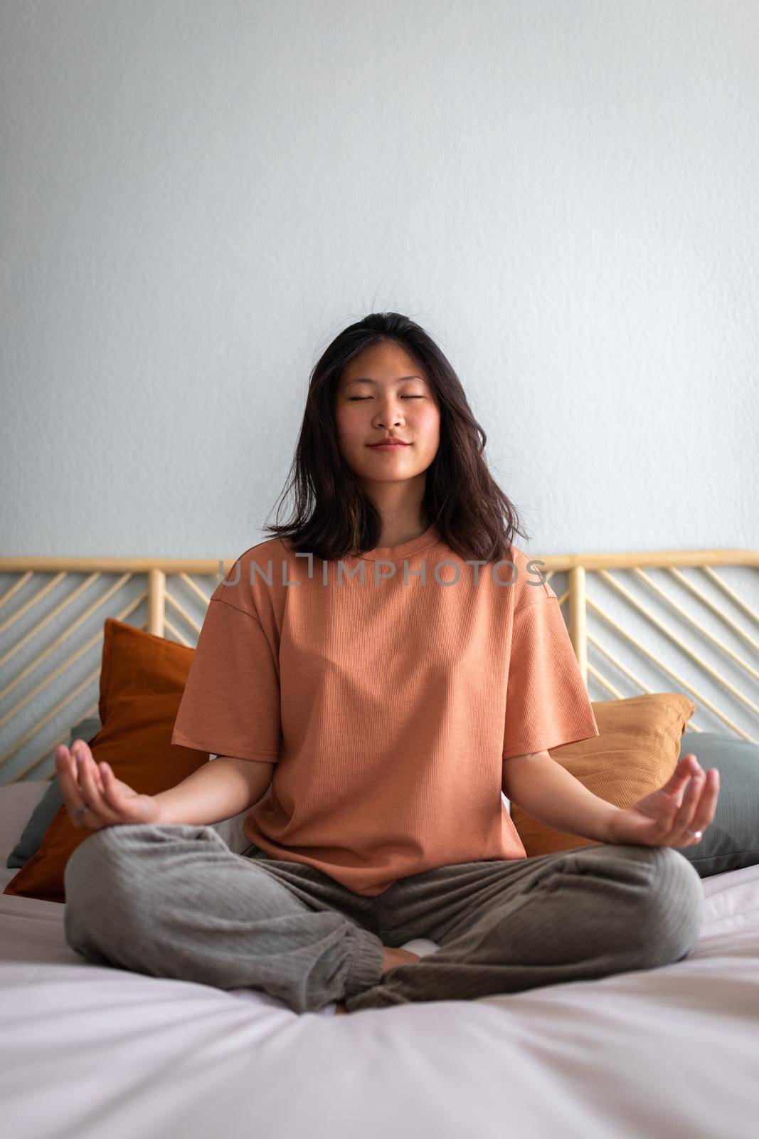 Front view vertical portrait of asian college student young woman meditating and doing breathing exercises on the bed in the morning to clear mind before day starts. Wellness and meditation concept.