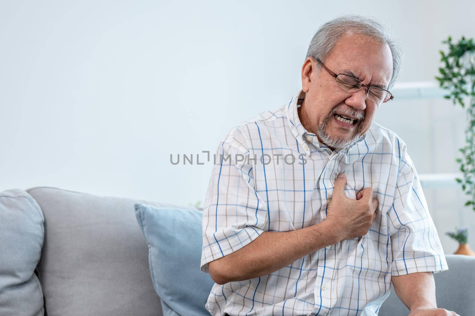 An agonizing senior man suffering from chest pain or heart attack alone in his living room. Serious health problem and feeling unwell concept for seniors.