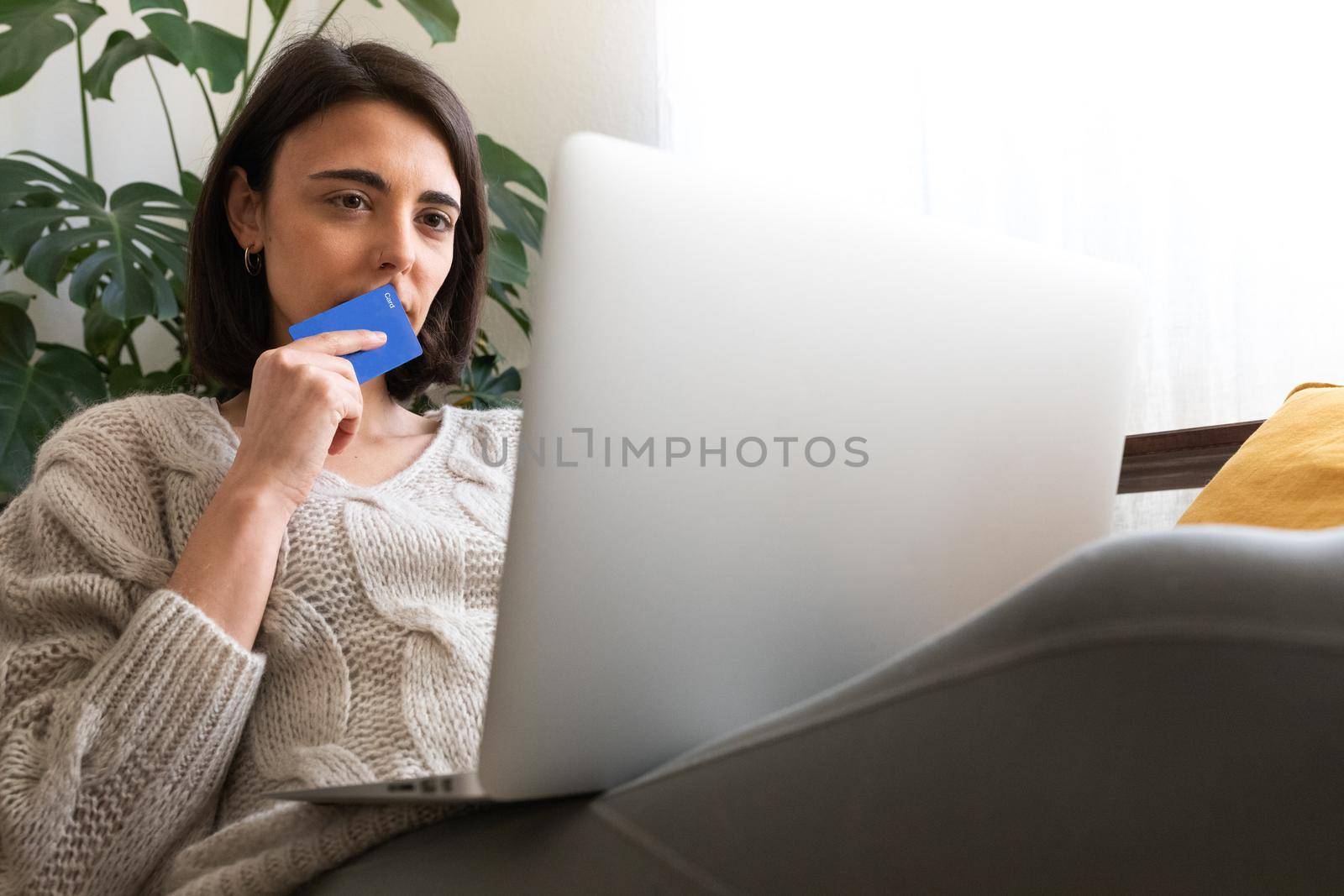 Pensive young caucasian woman in doubt holds credit card wondering if buying online. Female checking online bank account. Copy space.Technology and money concepts.