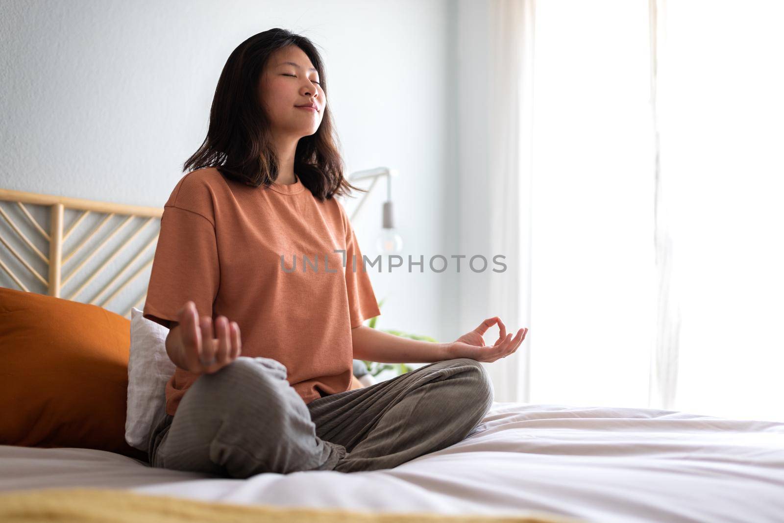 Young asian woman wearing comfortable clothes meditating on the bed in the morning to clear mind before day starts. Copy space. Wellness and meditation concept.