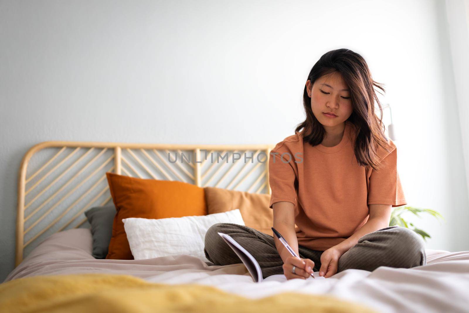 Pensive young Asian woman sitting on bed with serious expression writing on journal in cozy bedroom. Copy space. Lifestyle concept.