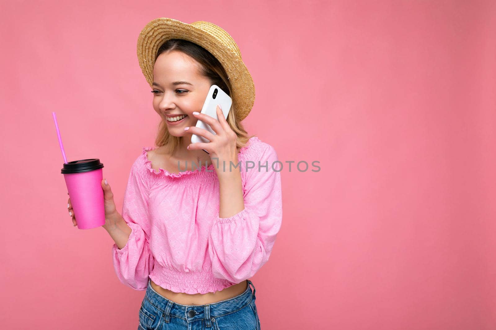Photo of beautiful positive emotional young blonde woman wearing pink crop blouse and straw hat talking on cellphone while drinking beverage isolated on pink background with copy space by TRMK