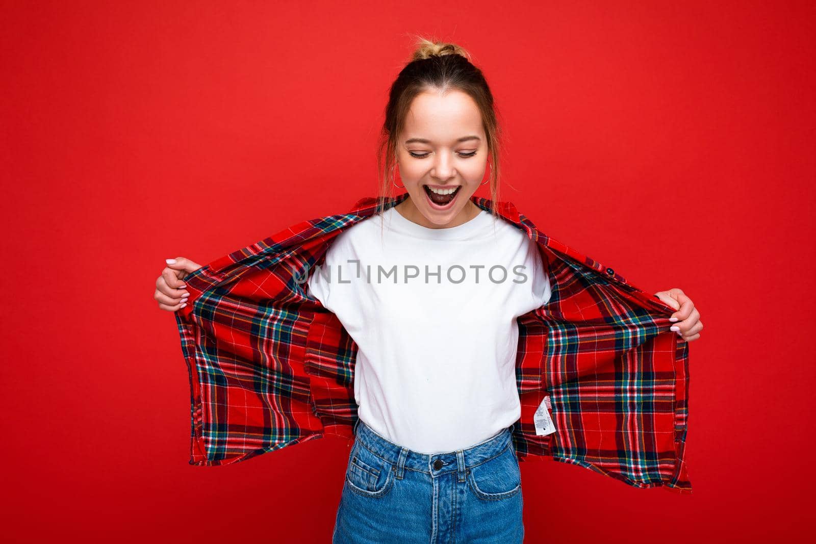 Young attractive surprised happy hipster blonde woman in stylish red shirt and casual white t-shirt. Sexy carefree female person posing near red wall in studio. Positive model with natural makeup. Copy space by TRMK