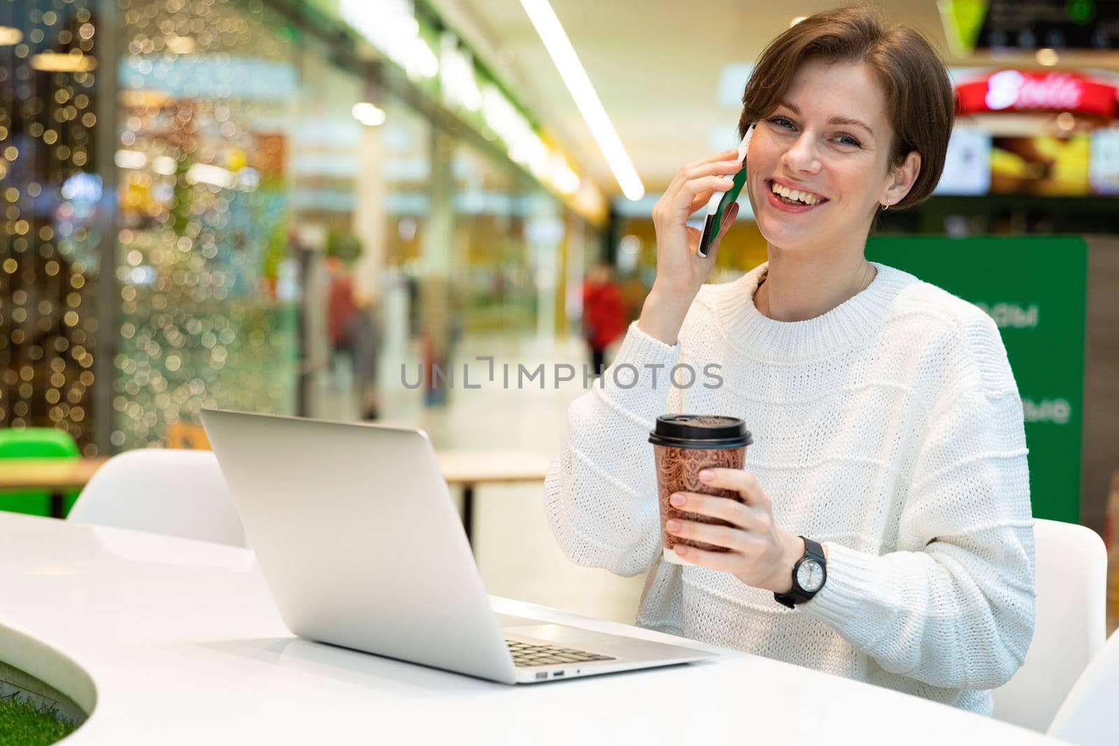 Young attractive positive smiling happy woman sitting at a shopping center at a table and working at a computer laptop, using mobile phone. Freelance and business concept.