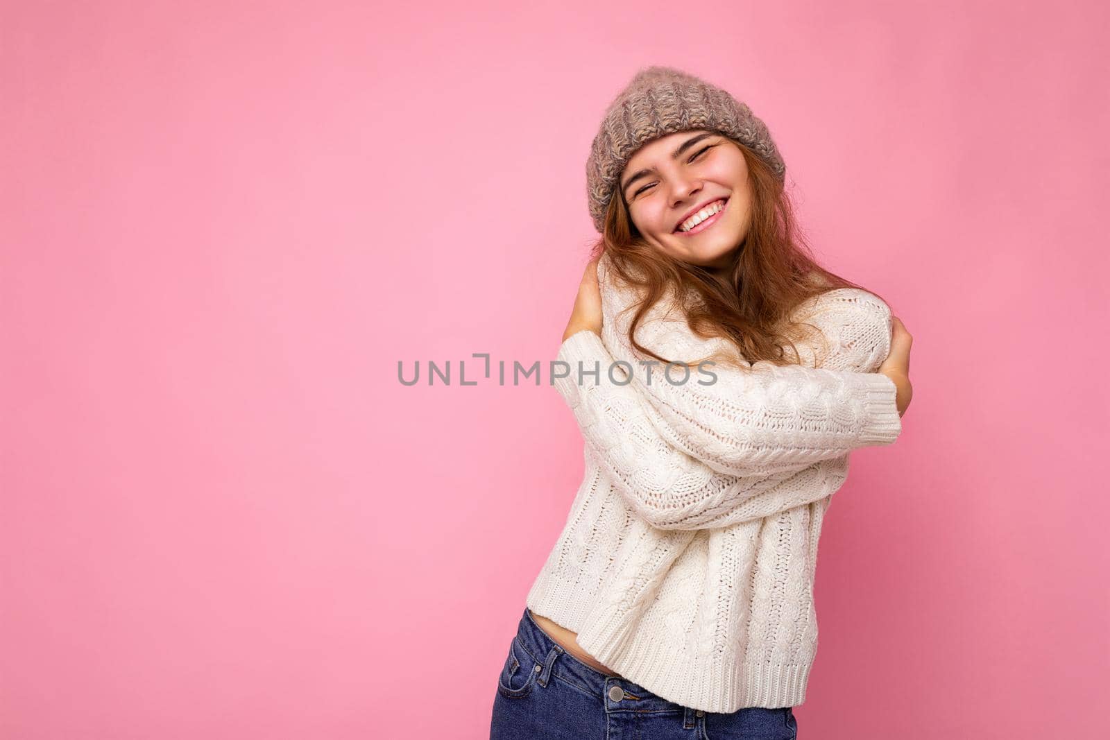 Shot of beautiful happy positive smiling young brunette woman isolated over pink background wall wearing knitted white sweater and grey warm hat and having fun. Free space