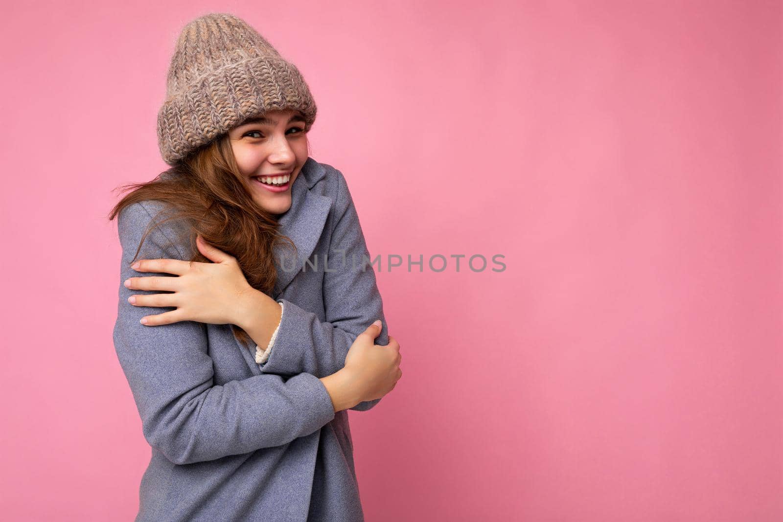 Shot of fascinating happy smiling young brunette woman isolated over pink background wall wearing grey autumn coat and grey warm hat looking at camera and feeling cold by TRMK