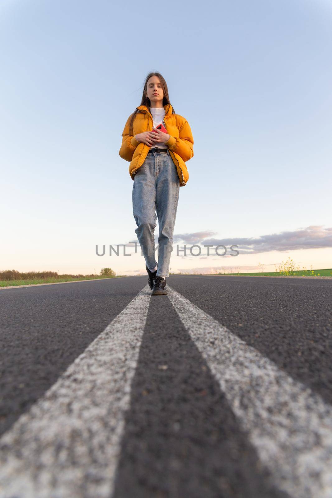 Cool modern teen girl poses on a lonely road while the sky background looks blue with clouds at sunset.