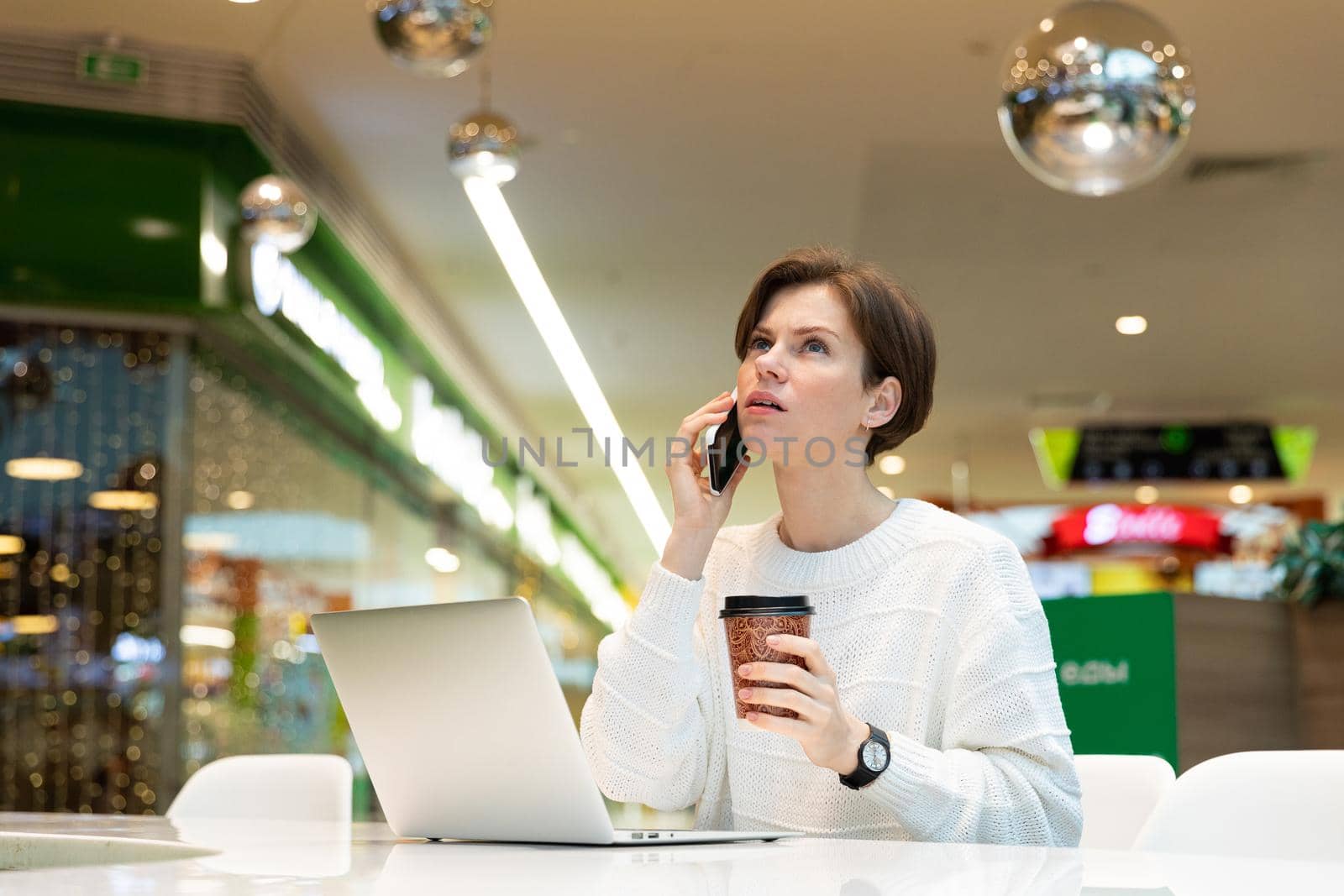 Young attractive woman sitting at a shopping center at a table and working at a computer laptop, using mobile smartphone. Cafe interior. Freelance and business concept.