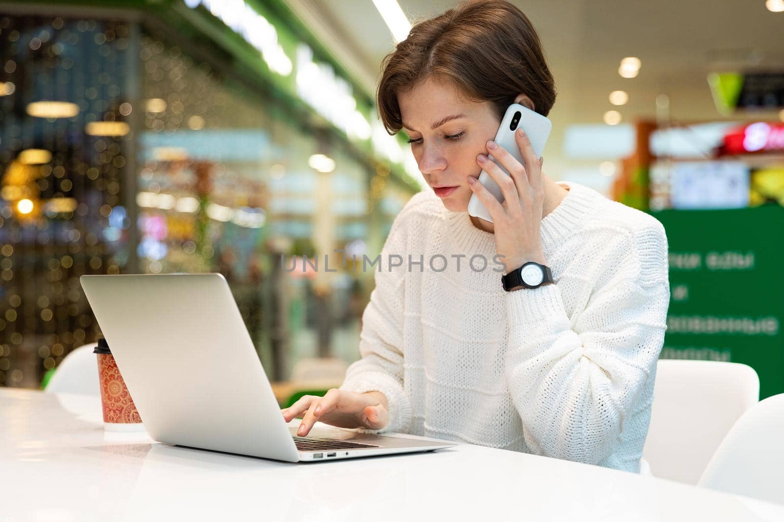 Young attractive woman sitting at a shopping center at a table and working at a computer laptop, using mobile phone. Cafe interior. Freelance and business concept.