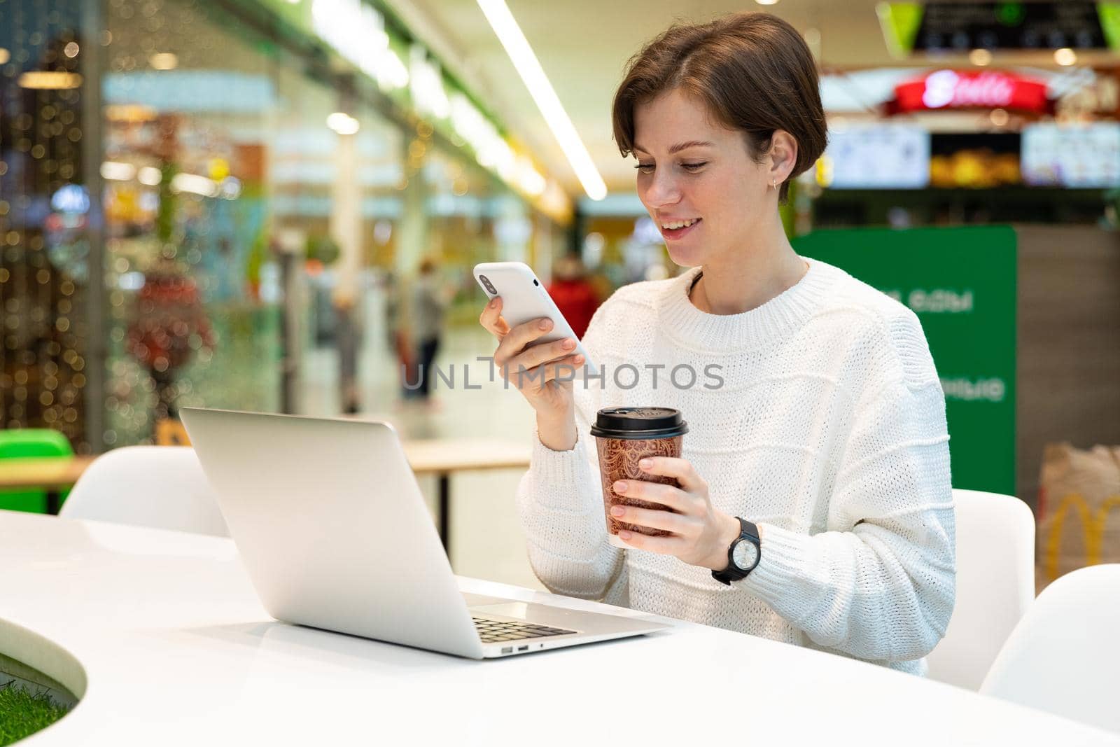 Young attractive woman sitting at a shopping center at a table and working at a computer laptop, using mobile phone. Freelance and business concept.