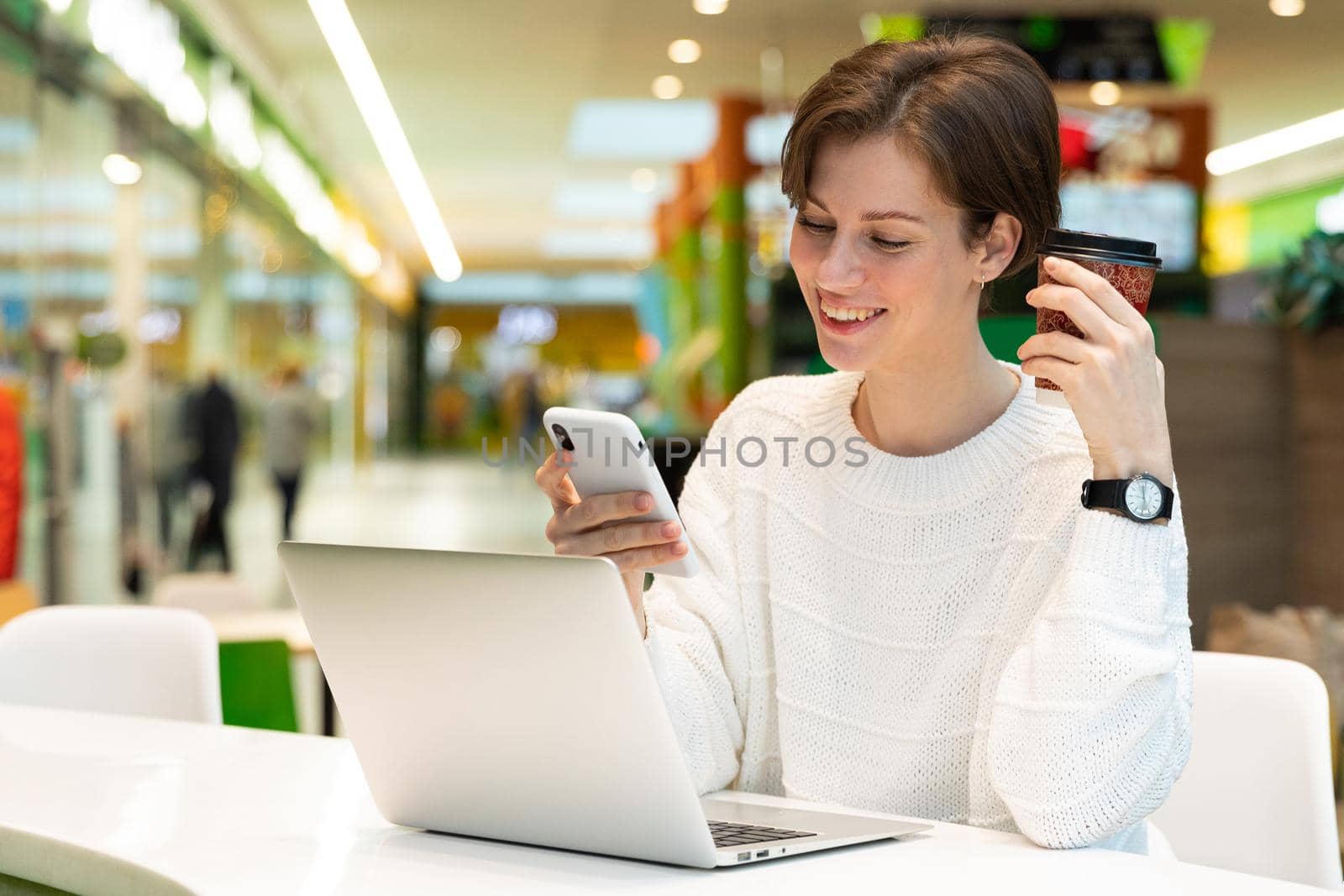 Young beautiful woman sitting at a shopping center at a table and working at a computer laptop, using mobile phone. Freelance and business concept by TRMK