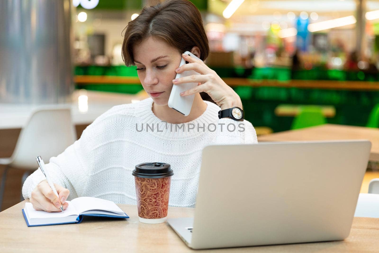 Young beautiful pretty brunette woman wearing white sweatshirt sitting at a shopping center at a table and working at a computer laptop, using mobile phone, writing information at notebook. Freelance and business concept.