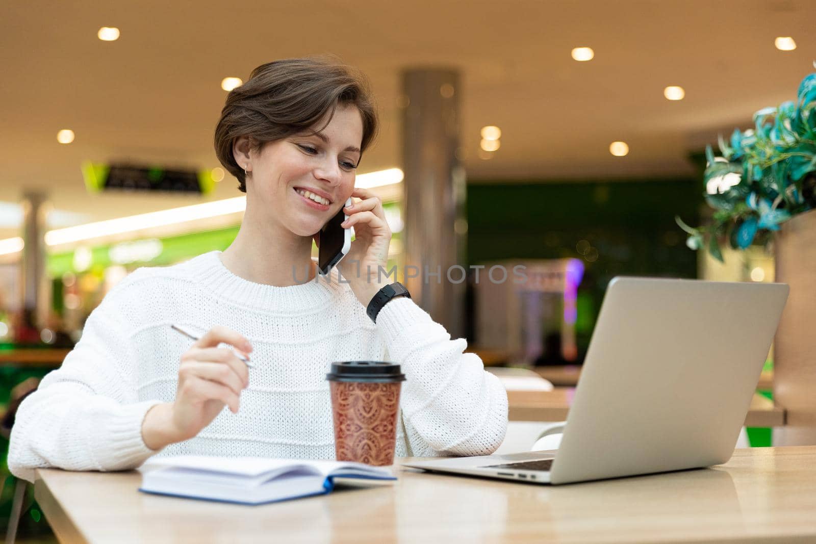 Young attractive brunette woman sitting at a shopping center at a table and working at a computer laptop, using mobile phone. Freelance and business concept.