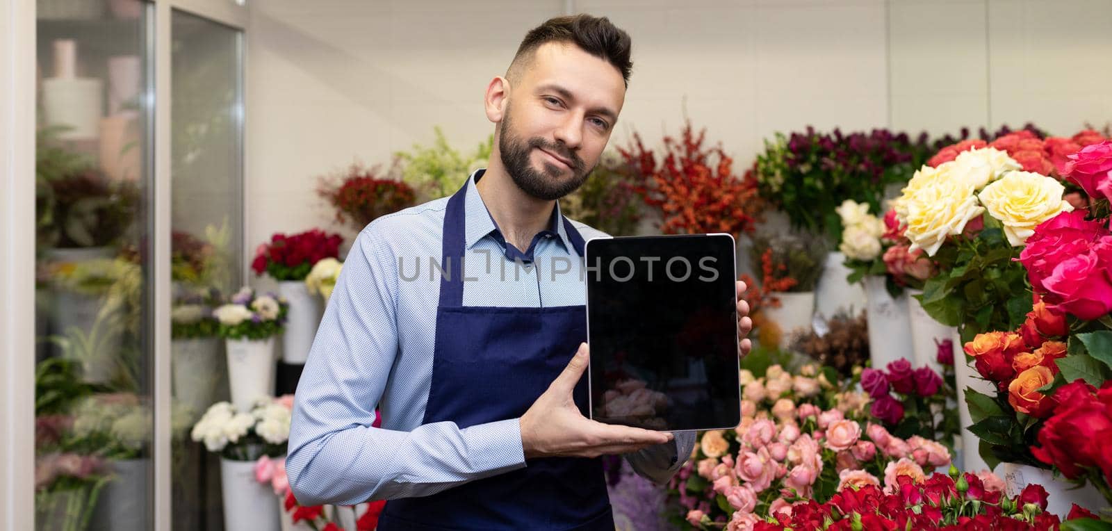 florist surrounded by fresh flowers demonstrates a tablet screen in a bouquets shop, concept of sale for Valentine's Day.