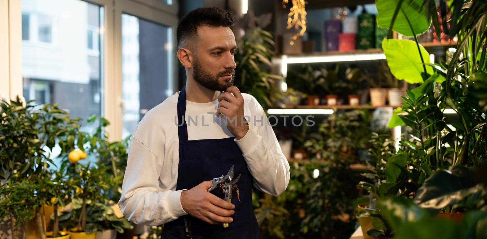 thoughtfully male florist with secateurs in hands among potted plants.