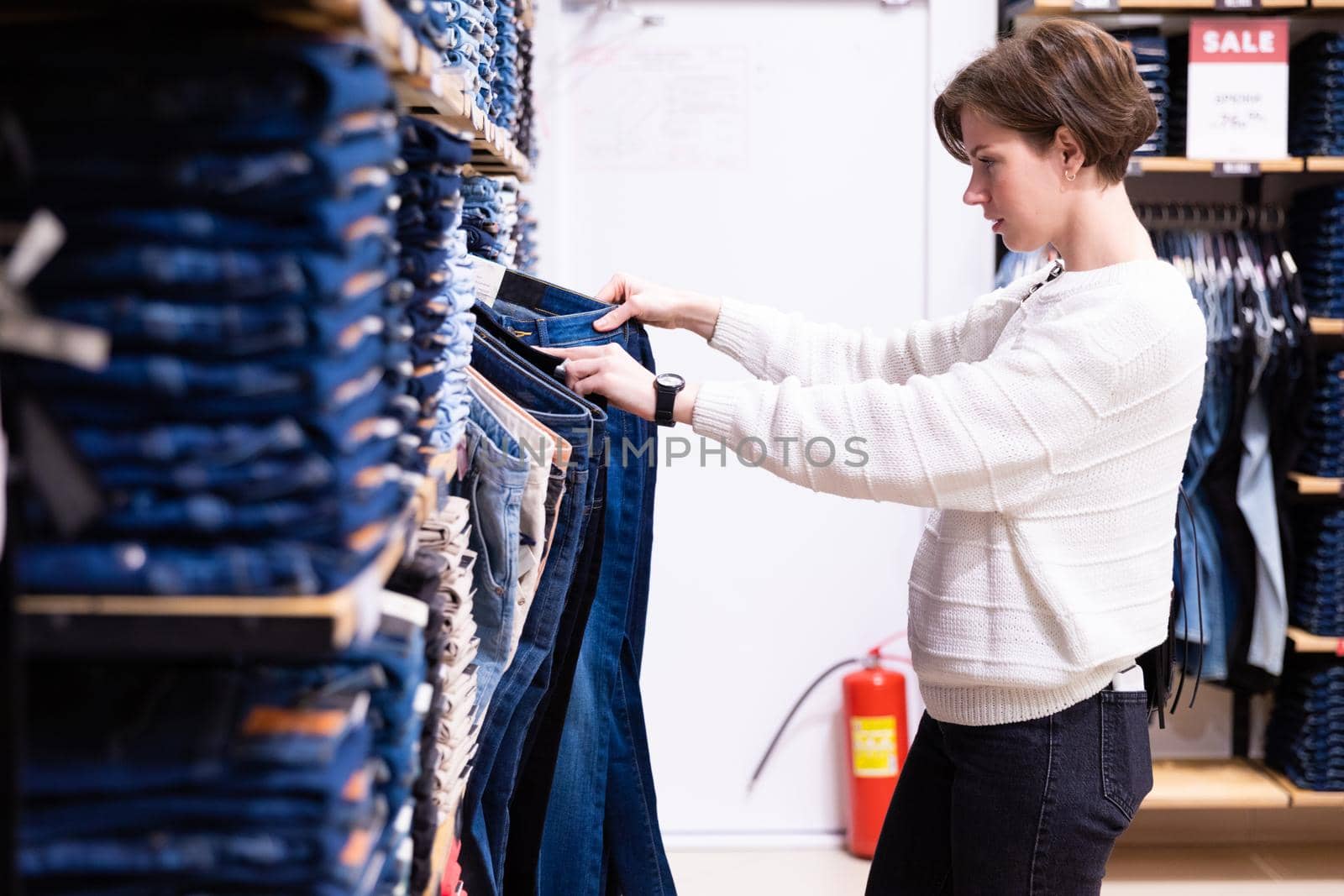 Photo of young attractive brunette woman with a short haircut in a white sweater chooses stylish and casual jeans and denim clothes in a store in a shopping centre by TRMK