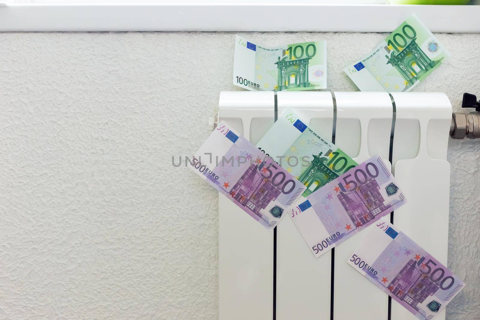 Paper banknotes of 100 and 500 euros on a small white radiator for heating the house. The concept of expensive heating costs and the energy crisis in Europe.