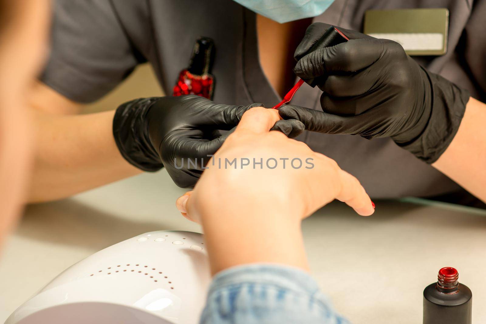 Manicure varnish painting. Close-up of a manicure master wearing rubber black gloves applying red varnish on a female fingernail in the beauty salon. by okskukuruza