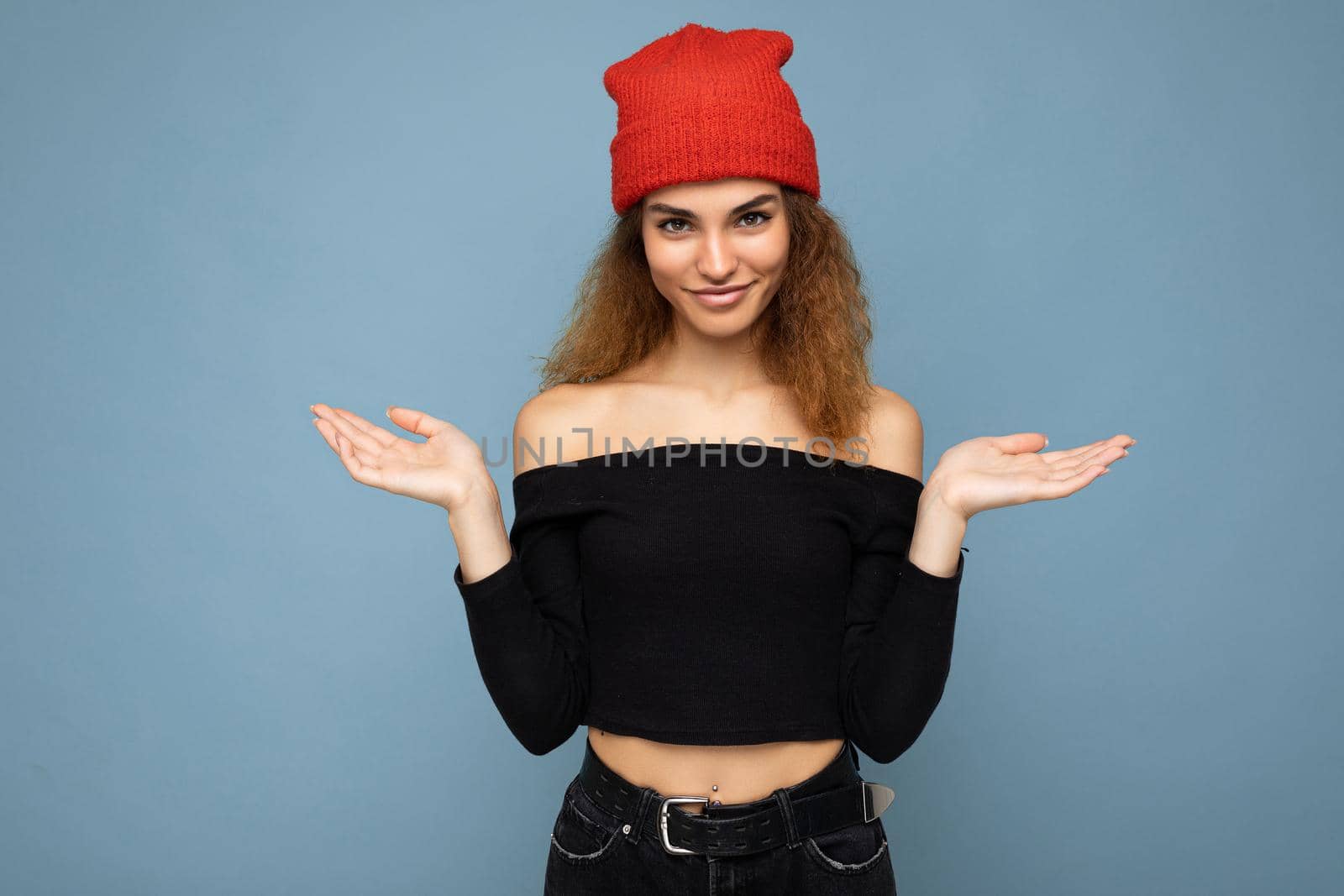 Photo of young positive happy attractive brunette curly woman with sincere emotions wearing black crop top and red hat isolated on blue background with empty space and having doubts.