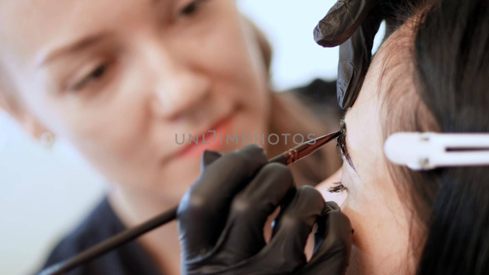 close up. young woman gets eyebrow correction procedure. kosmetolog- makeup artist in black rubber gloves applies paint with brush on eyebrows in beauty saloon. Professional care for face. High quality photo