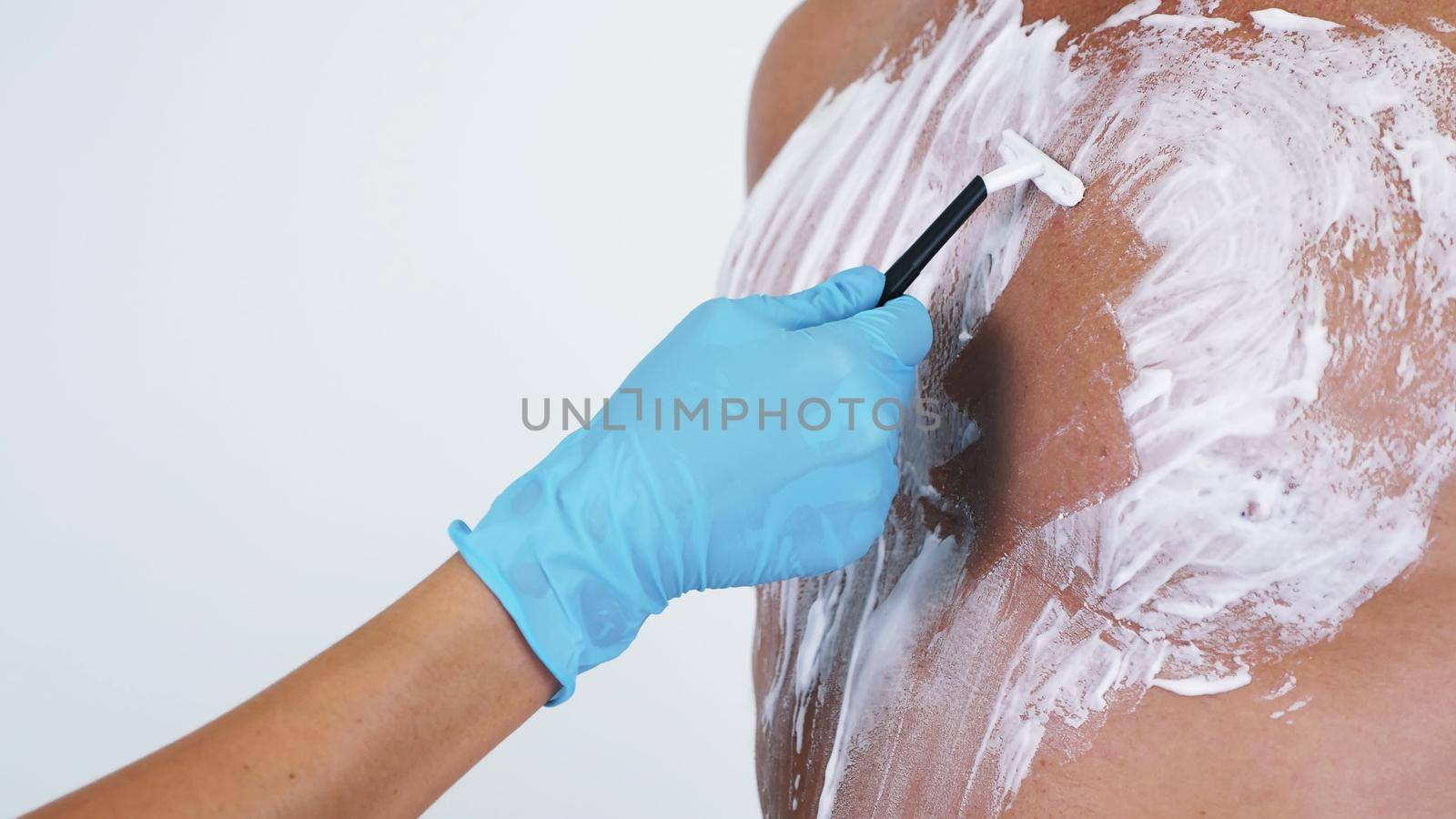 Close-up, cosmetologist female hands in gloves extrude white shaving foam from the tube and spread it on Naked hairy male Torso, chest. on white background. High quality photo