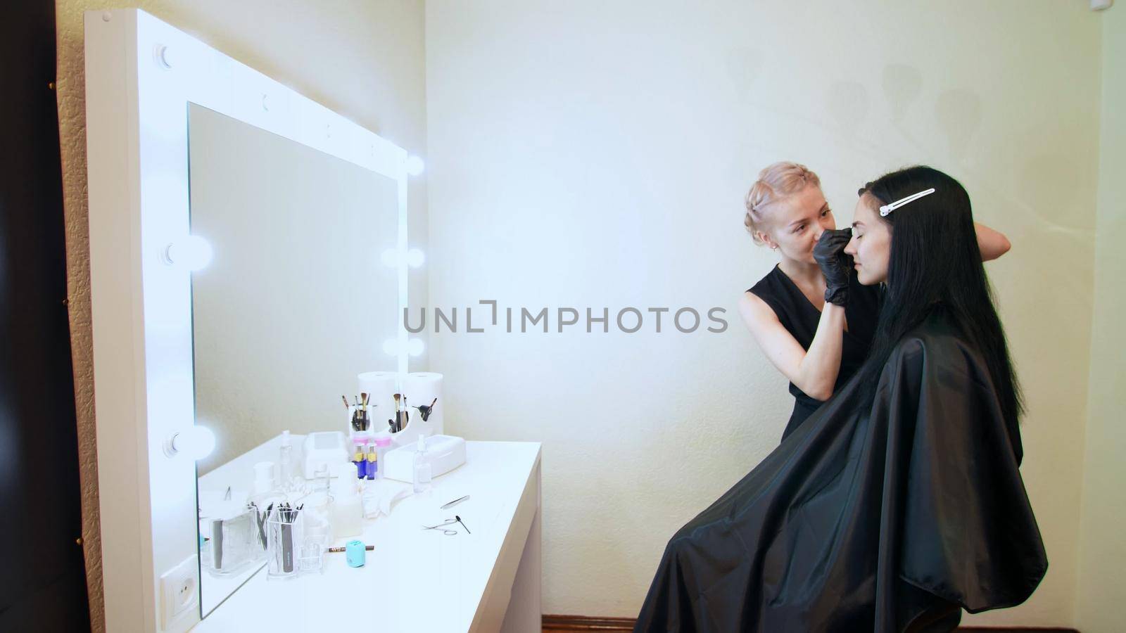 beauty saloon. the cosmetician in black rubber gloves hold tweezers and pull out eyebrows. Master corrects the shape of the eyebrows. near a large mirror with illumination. High quality photo