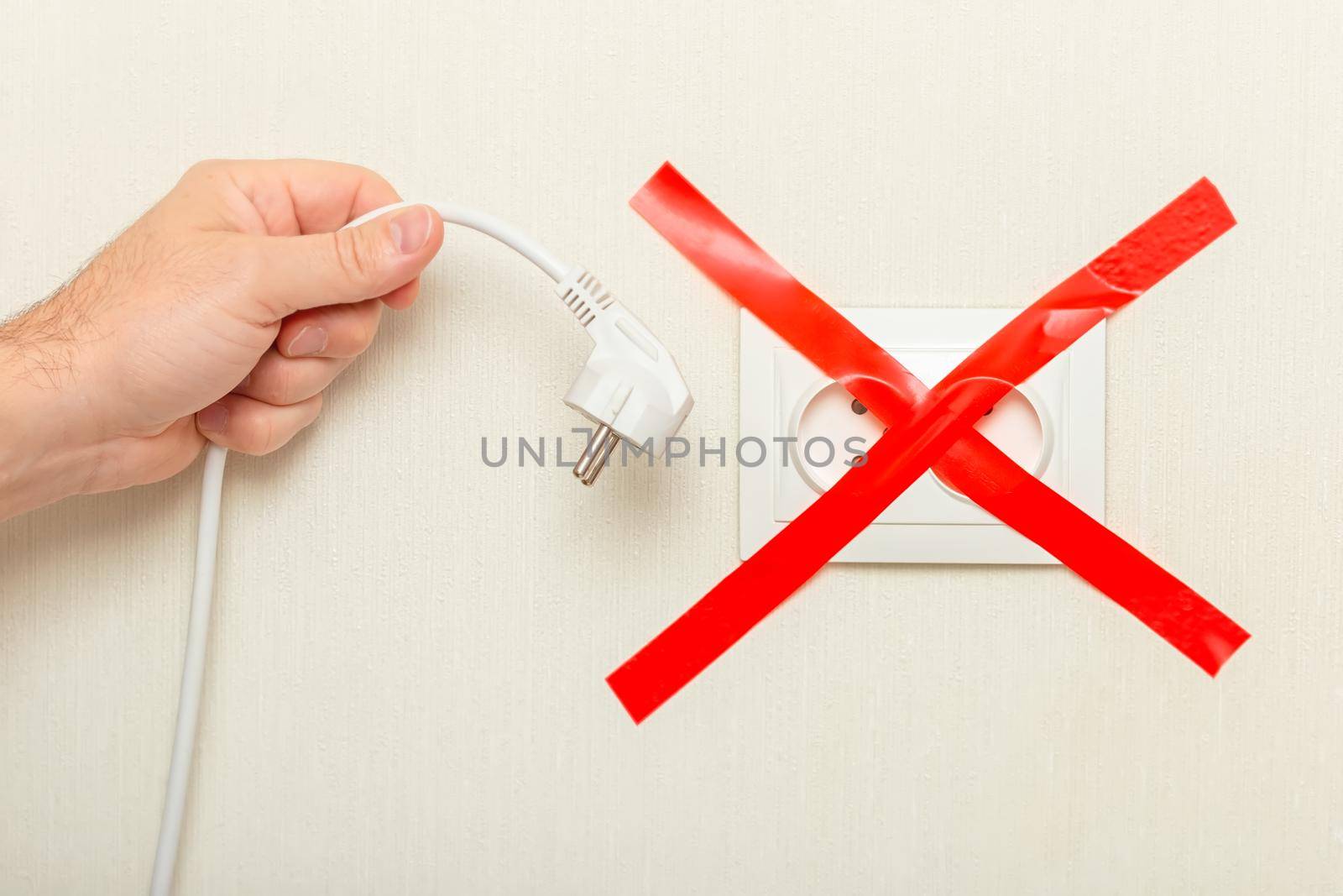 A mans hand holds a drooping white cord with a plug near an electrical outlet, tied with a red ribbon crosswise. The concept of the energy crisis in Europe